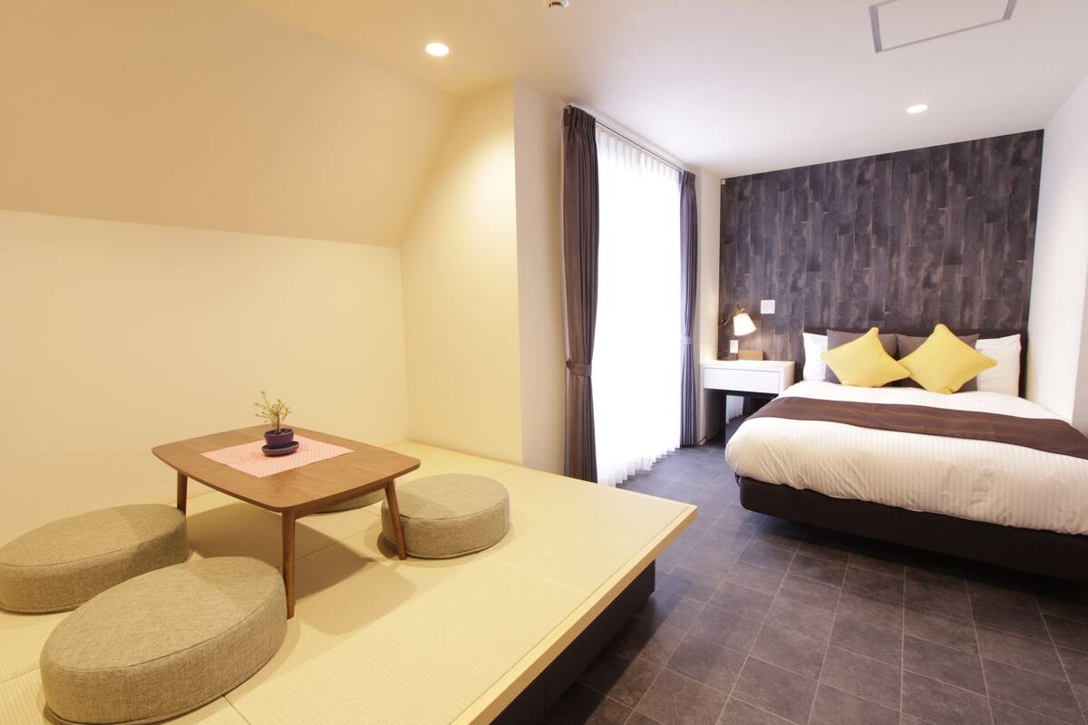 A modern hotel room with a large bed, a bedside table with a lamp, and a small seating area with cushions.
