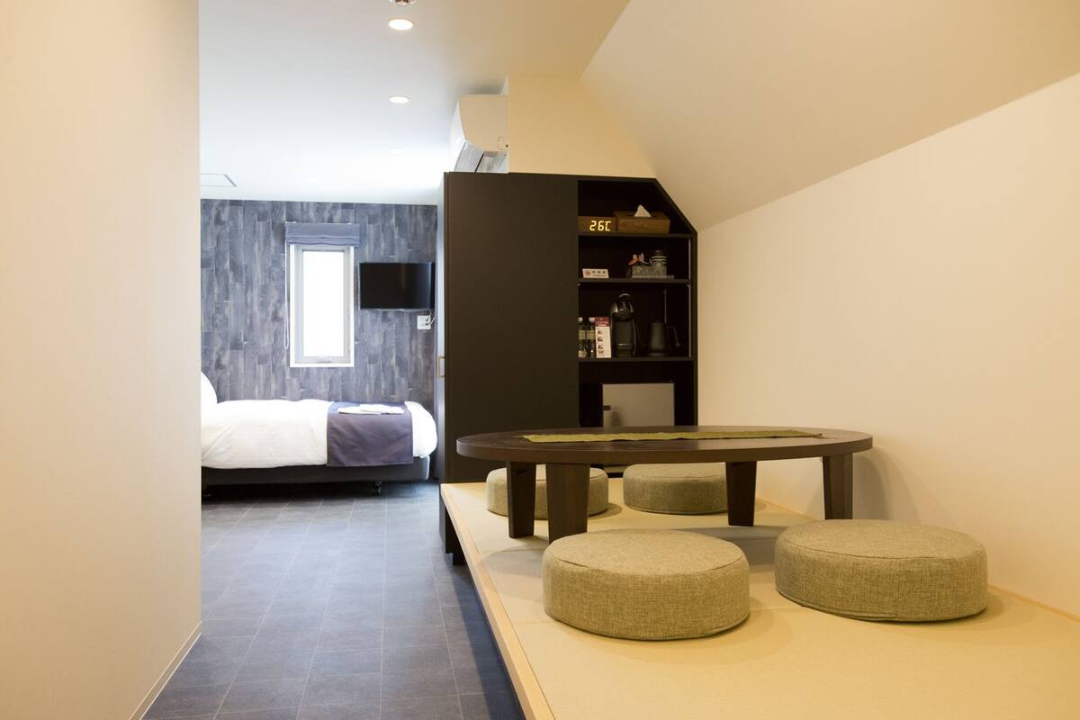 A modern hotel room with a bed, a small dining table, and ottomans.