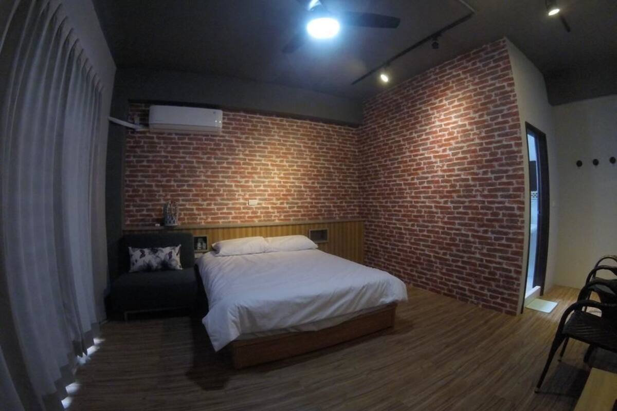 A bedroom with a brick wall, a bed, a nightstand, a chair, and a ceiling fan.