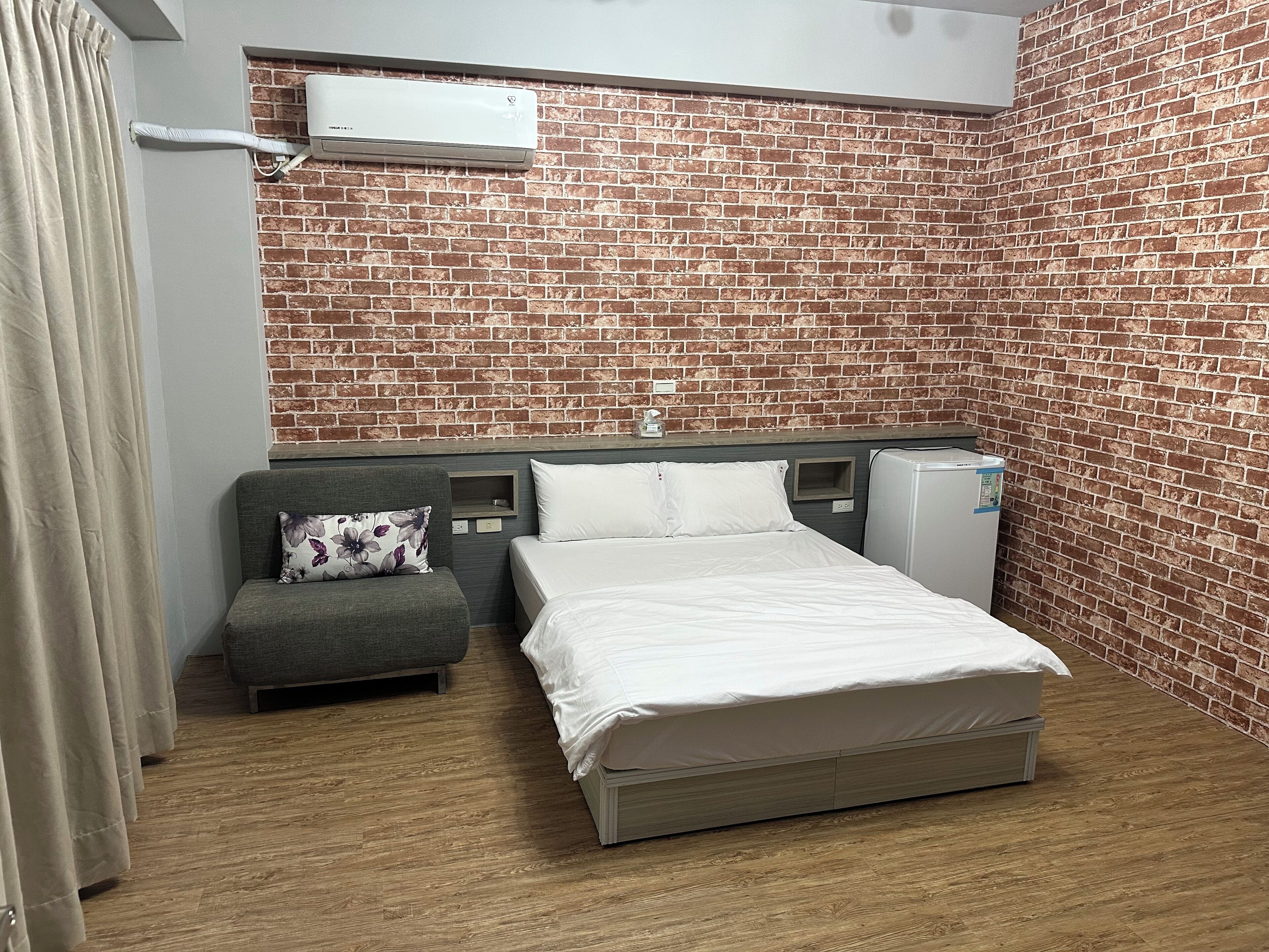 Een hotelkamer met een muur in baksteenmotief, een bed met witte beddengoed, een grijze bank met een bloemenkussen, en een kleine witte koelkast.