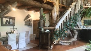 Un intérieur rustique avec un escalier en bois, un coin salon confortable avec des chaises blanches, un meuble en bois et un grand tronc d’arbre.