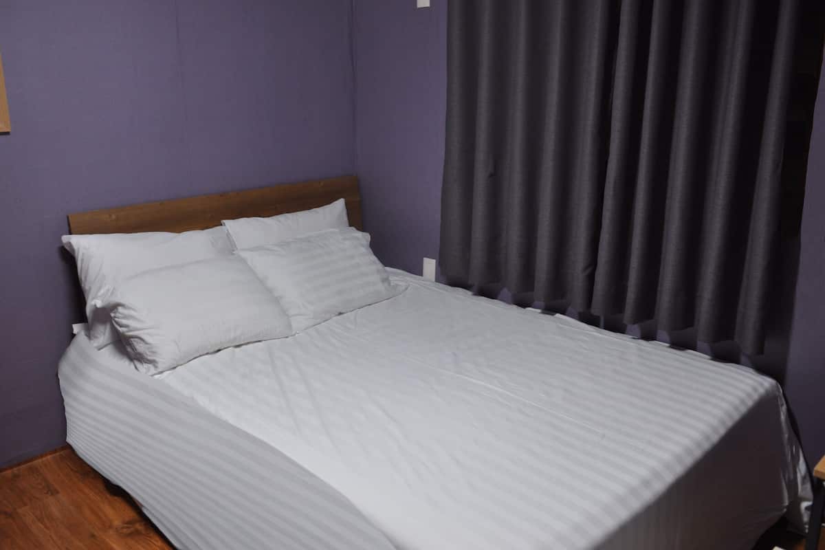 A neatly made bed with a wooden headboard, a purple wall, and a dark curtain.