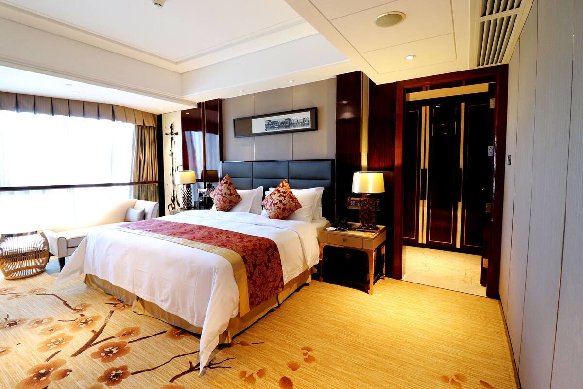 A hotel room with a large bed, a nightstand with a lamp, a wooden floor, and a large window with curtains.