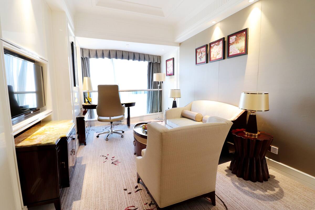 A hotel room with a TV, a sofa, a desk with a lamp, a chair, and a decorative side table.