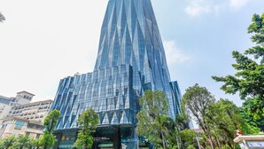 Un rascacielos alto y moderno con fachada de vidrio geométrica singular, rodeado de árboles y otros edificios.