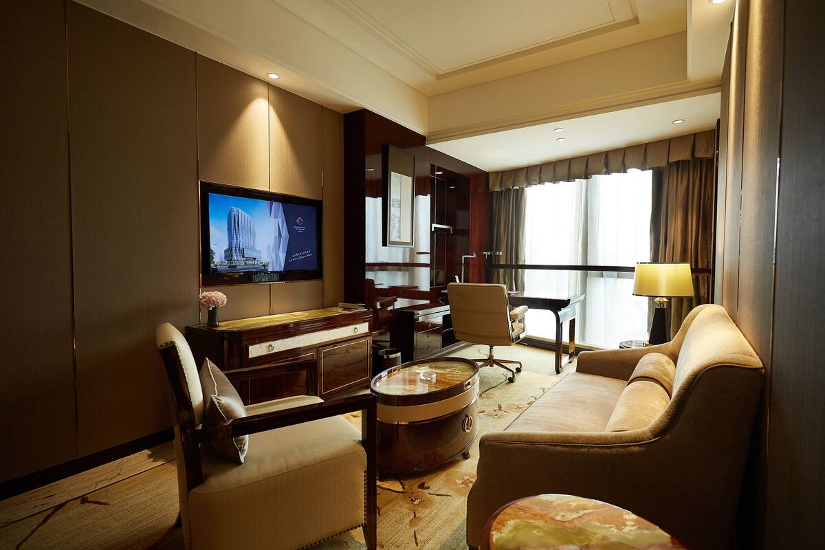 A hotel room with a flat-screen TV, a sofa, a chair, a small round coffee table, and a desk with a lamp.