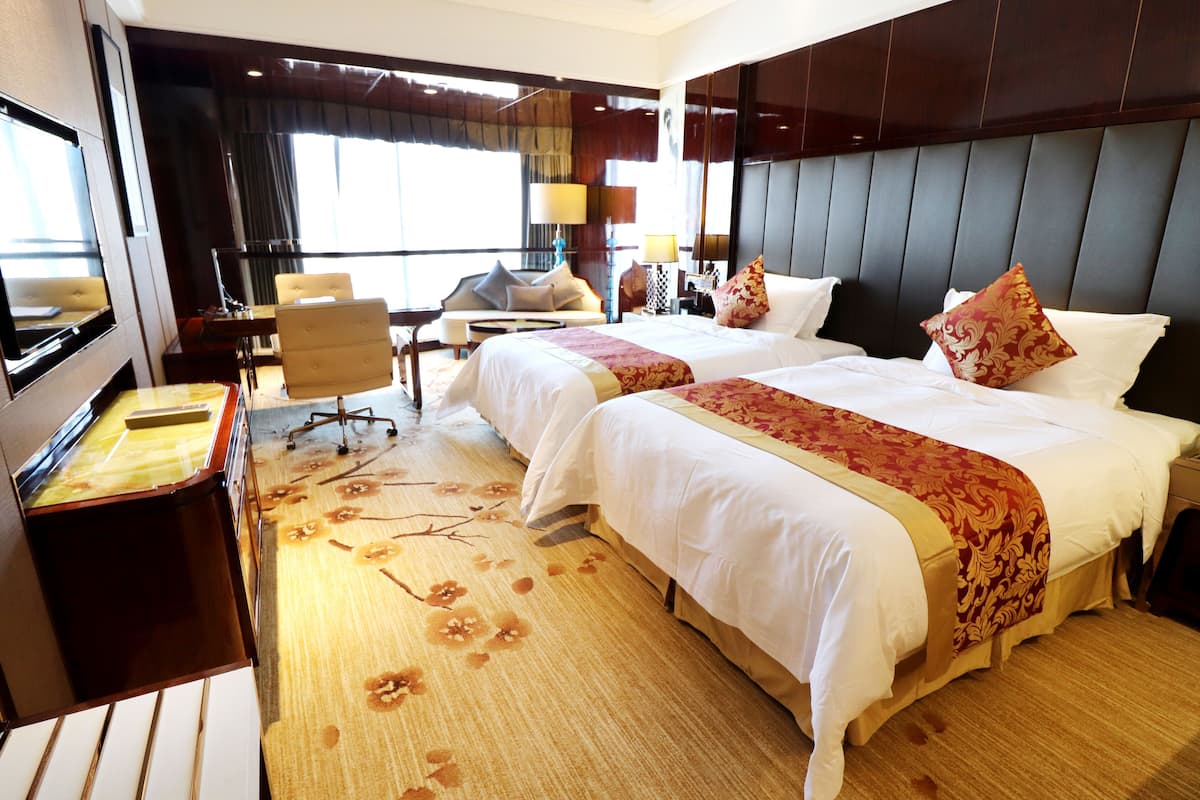 A hotel room with two beds, a desk, a chair, a TV, and a large window.