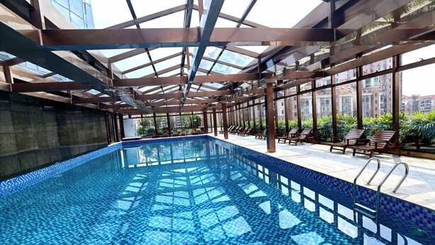Piscina interior con suelo de baldosas y techo de cristal, rodeada de sillones de descanso y vistas a edificios.