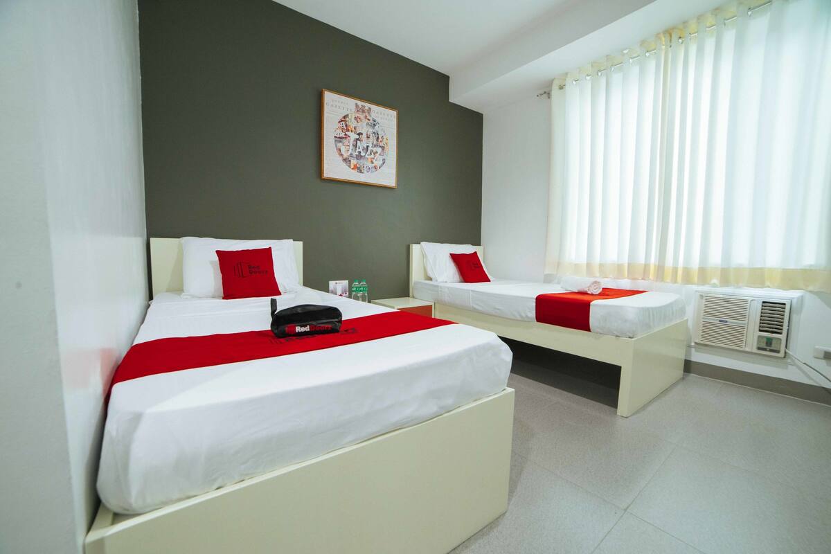 A hotel room with two beds, each with white bedding and red accents, a wall-mounted artwork, and a window with curtains.