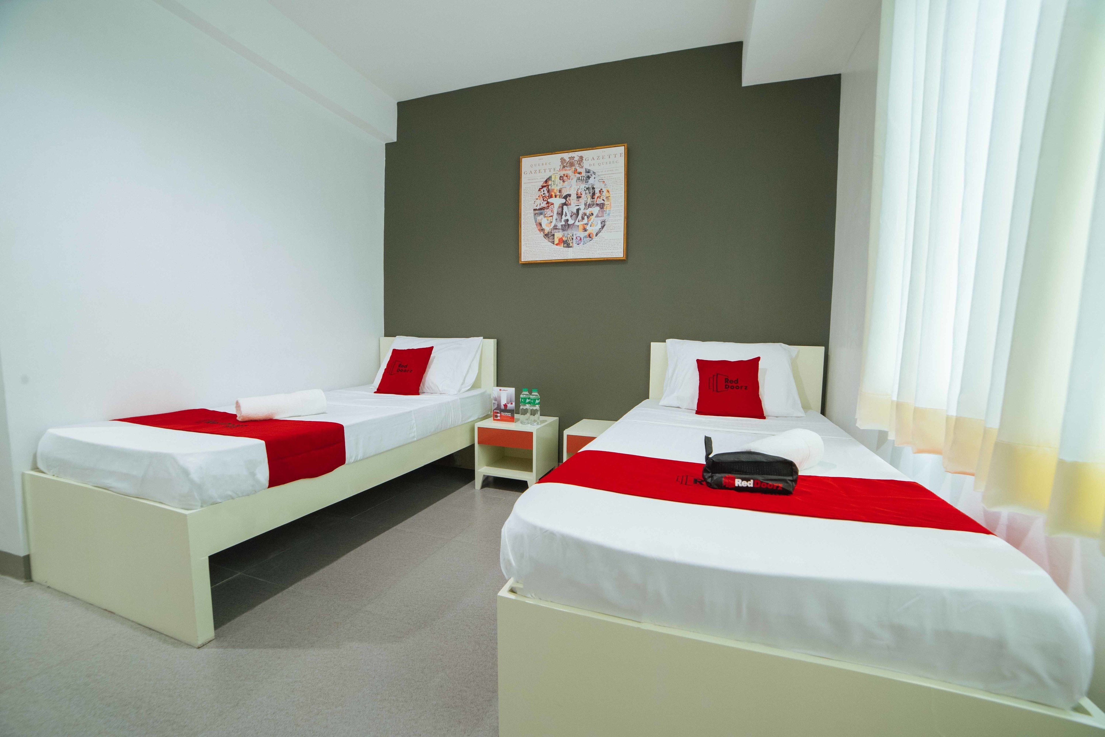 Ein Hotelzimmer mit zwei Betten, jeweils mit weißer Bettwäsche und roten Akzenten, einem kleinen Nachttisch mit einer Flasche und einem gerahmten Bild an der Wand.