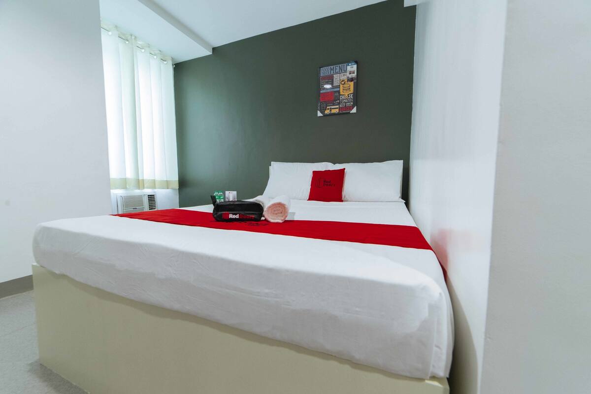 A neatly made bed with white bedding and a red accent pillow, a wall-mounted menu board, and a window with curtains.