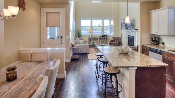 A modern kitchen with a large island, bar stools, and a dining area with a wooden table and chairs. - Whale of a Tale - Oyhut Bay Seaside Village (Ocean Shores)