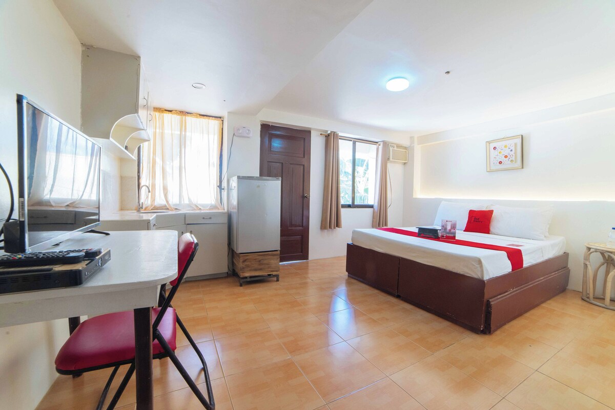 A compact room with a bed, a desk, a chair, a refrigerator, and a window with curtains.