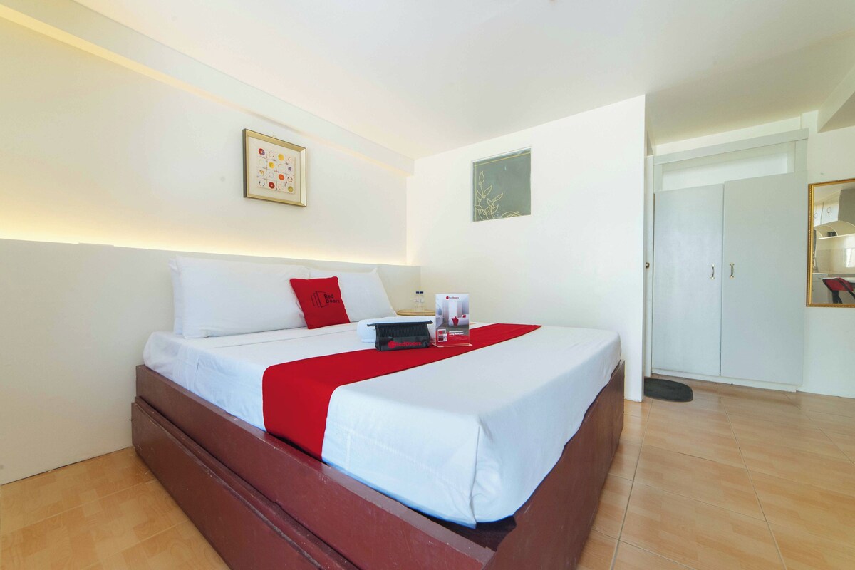 A neatly made bed with white bedding and a red accent, a framed picture on the wall, and a wardrobe in the background.