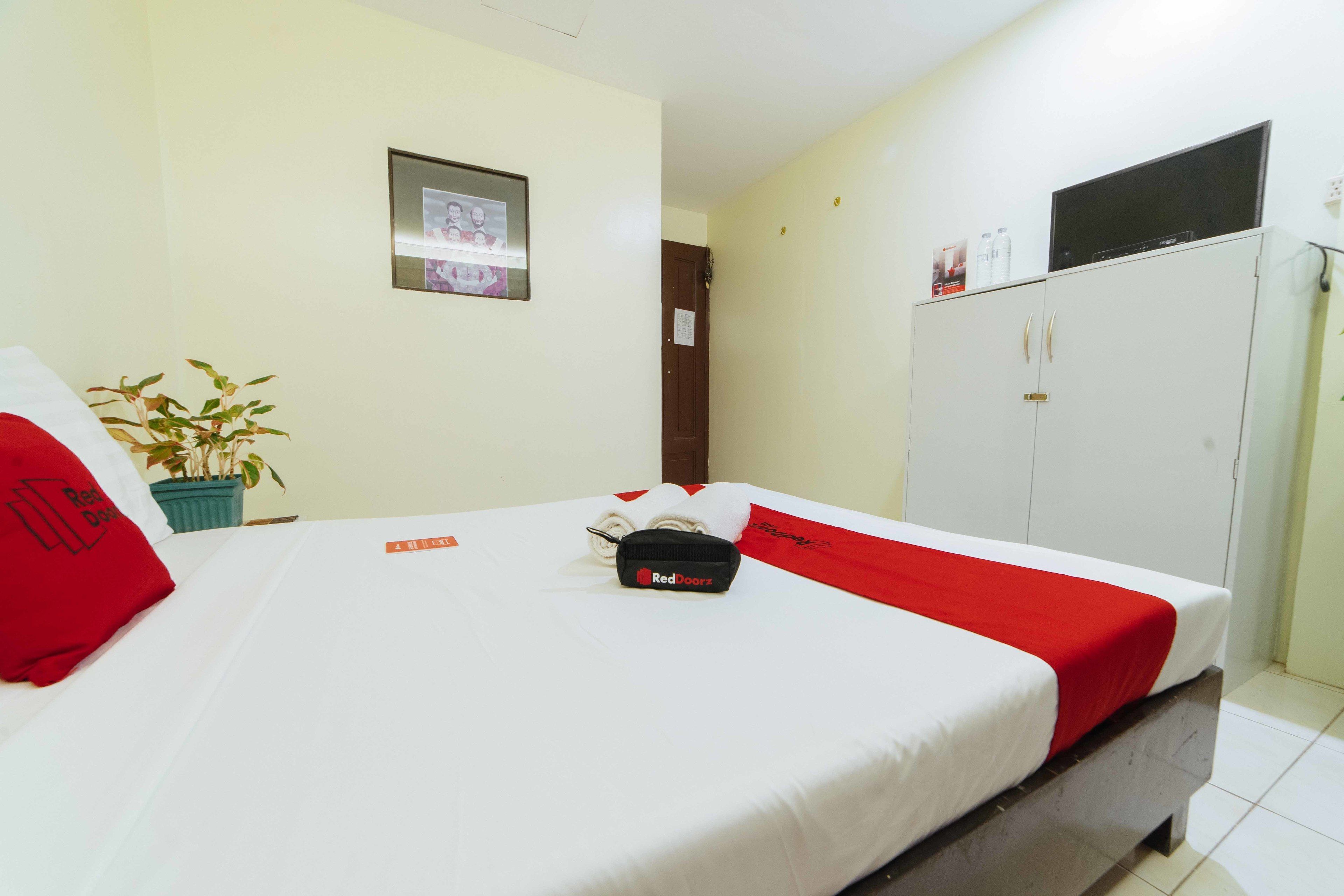 Quarto de hotel com cama, planta em vaso, guarda-roupa e televisão em cima do guarda-roupa.