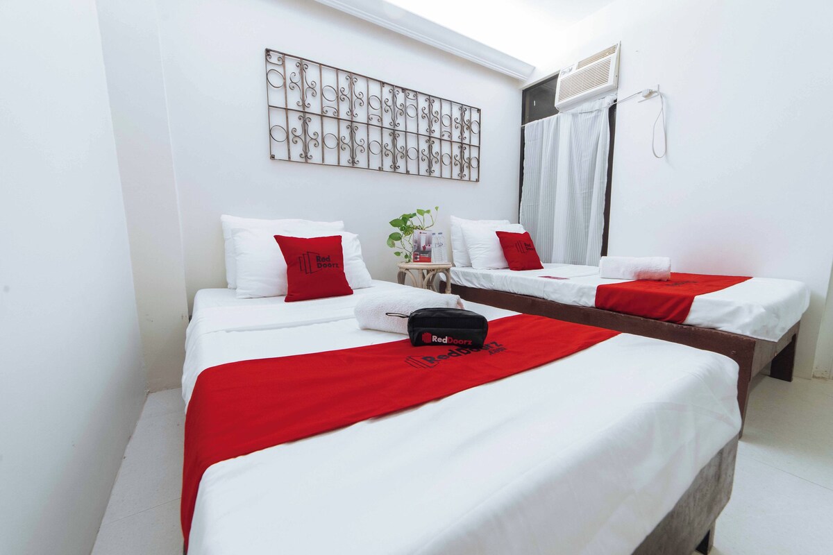 A hotel room with two single beds, each with white bedding and red accents, a small table with a plant, and a decorative metal piece on the wall.
