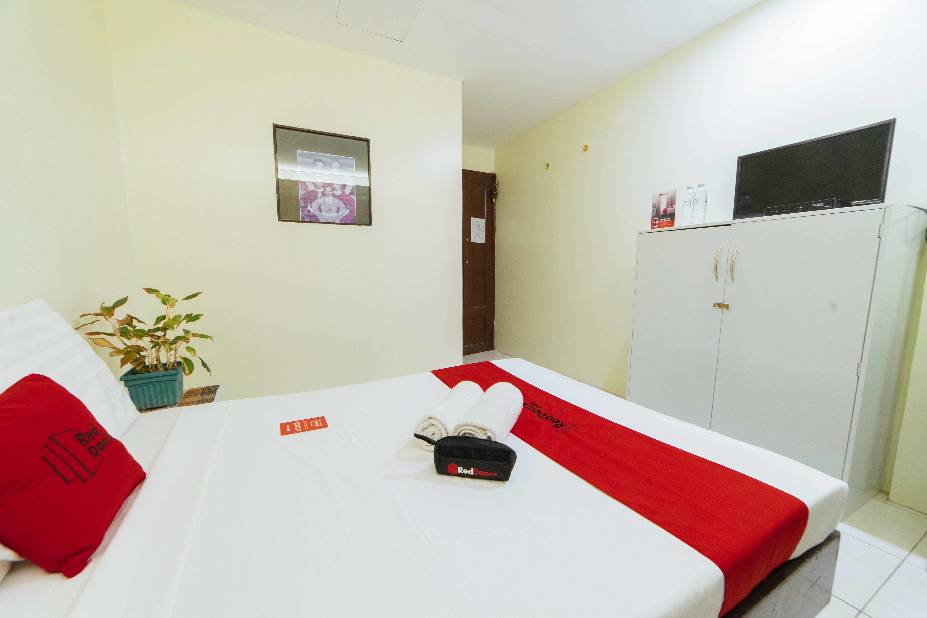 A hotel room with a bed, a red and white bedspread, a red pillow, a small potted plant, a television on top of a cabinet, and a framed picture on the wall.