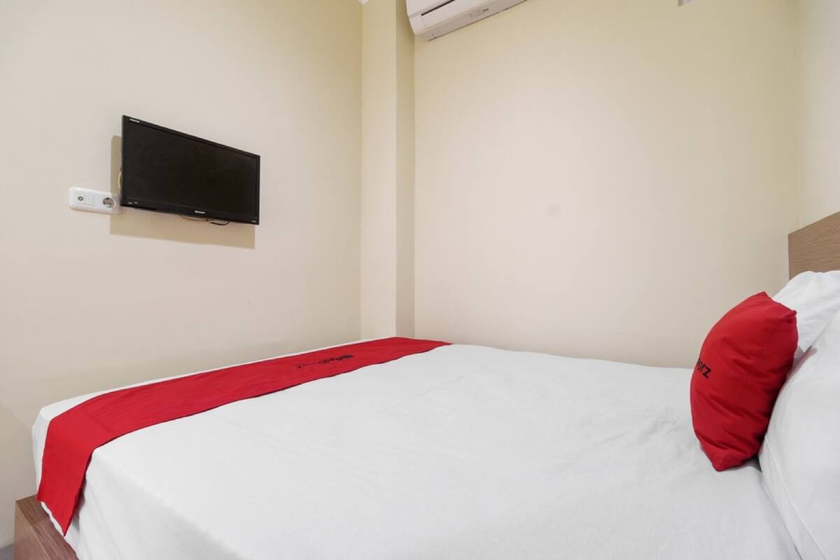 A single bed with a white and red bedspread, a red pillow, and a mounted flat-screen TV on the wall.
