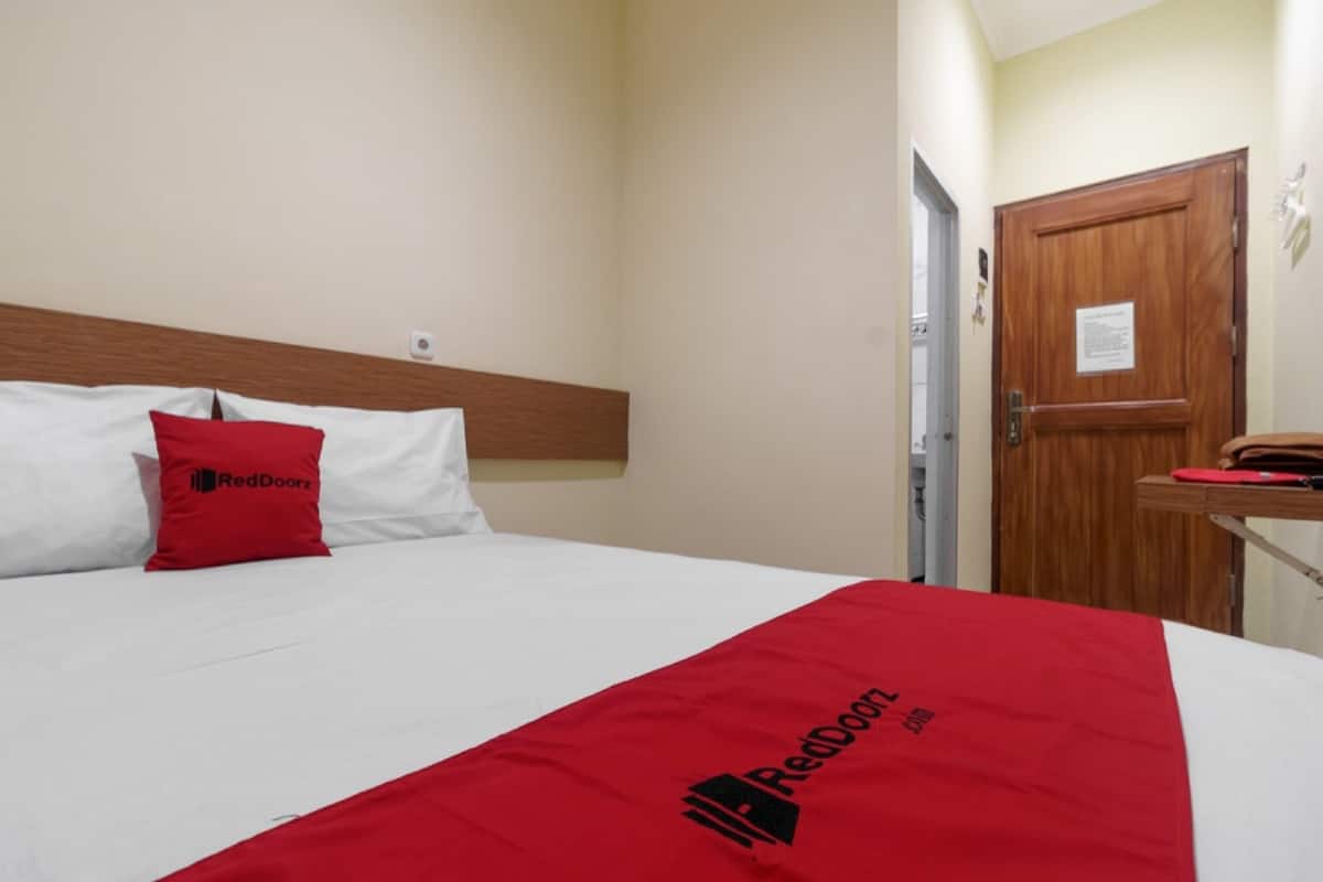 A hotel room with a bed, a wooden headboard, a red and white bedspread, a red pillow, and a wooden door.