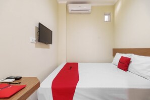 Une chambre d’hôtel avec un lit, un téléviseur fixé au mur, un climatiseur, une petite fenêtre et une table de chevet en bois avec un coussin rouge et un sac rouge.
