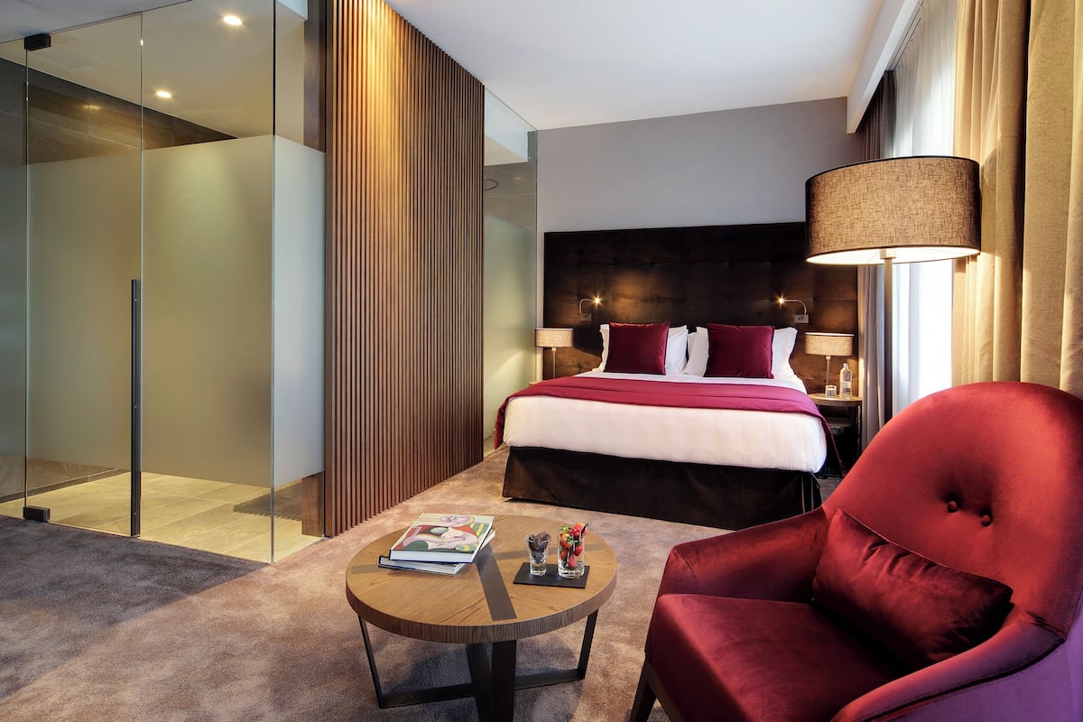 Una camera d'albergo moderna con un letto grande, una poltrona rossa, un tavolino rotondo con una rivista e un bicchiere d'acqua, e un box doccia in vetro.