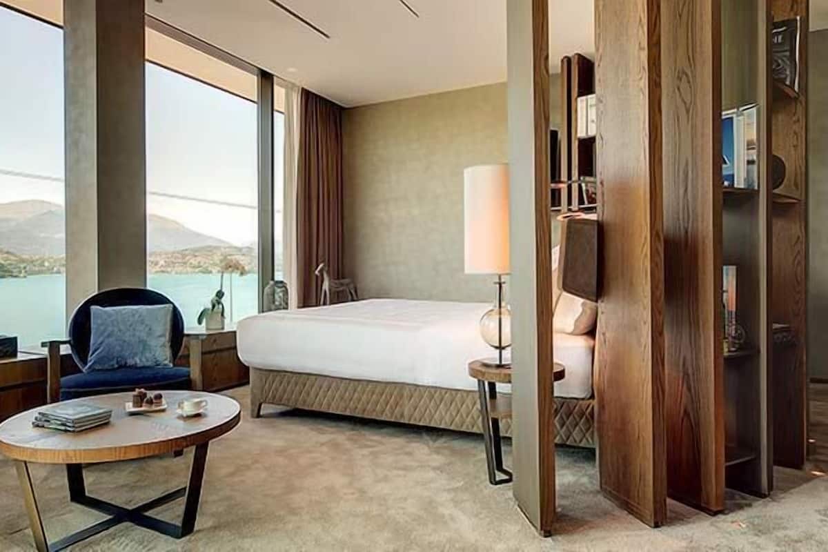 Una camera d'albergo con un letto, una scrivania, una sedia, una lampada e vista su un lago e delle montagne.