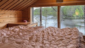 A bedroom with a large bed, wooden walls, a window overlooking a lake, and a small table with a bowl. - Sompsanniemi by Interhome (Kuusamo)