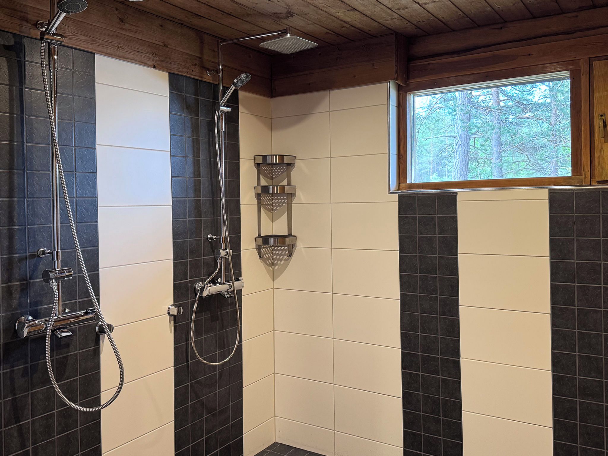 A shower with a handheld showerhead and a wall-mounted showerhead, a window with a view of trees, and a wooden ceiling.