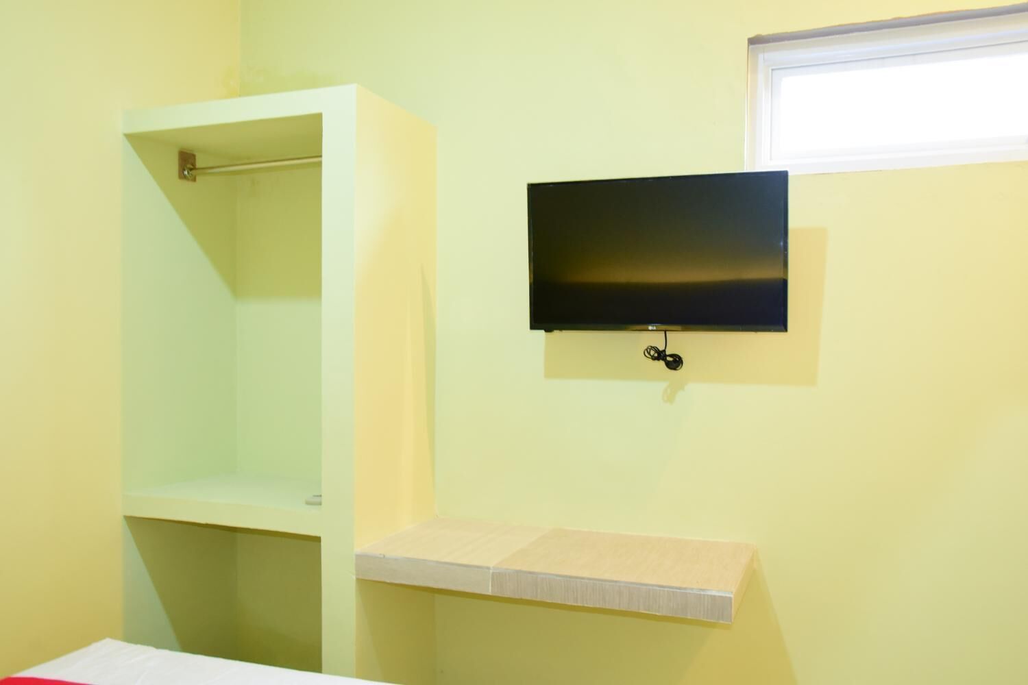 A room with a bed, a small shelf, a television mounted on the wall, and a window.