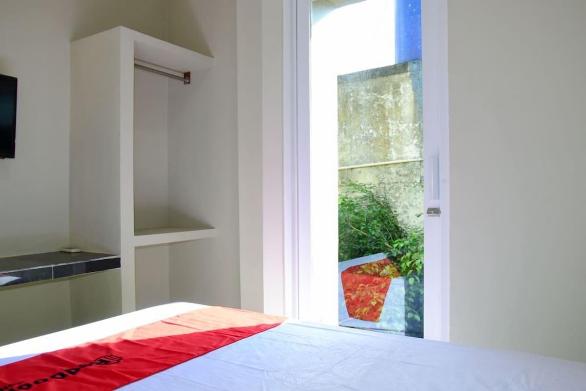 A hotel room with a bed, a small shelf, and a view of greenery through a glass door.