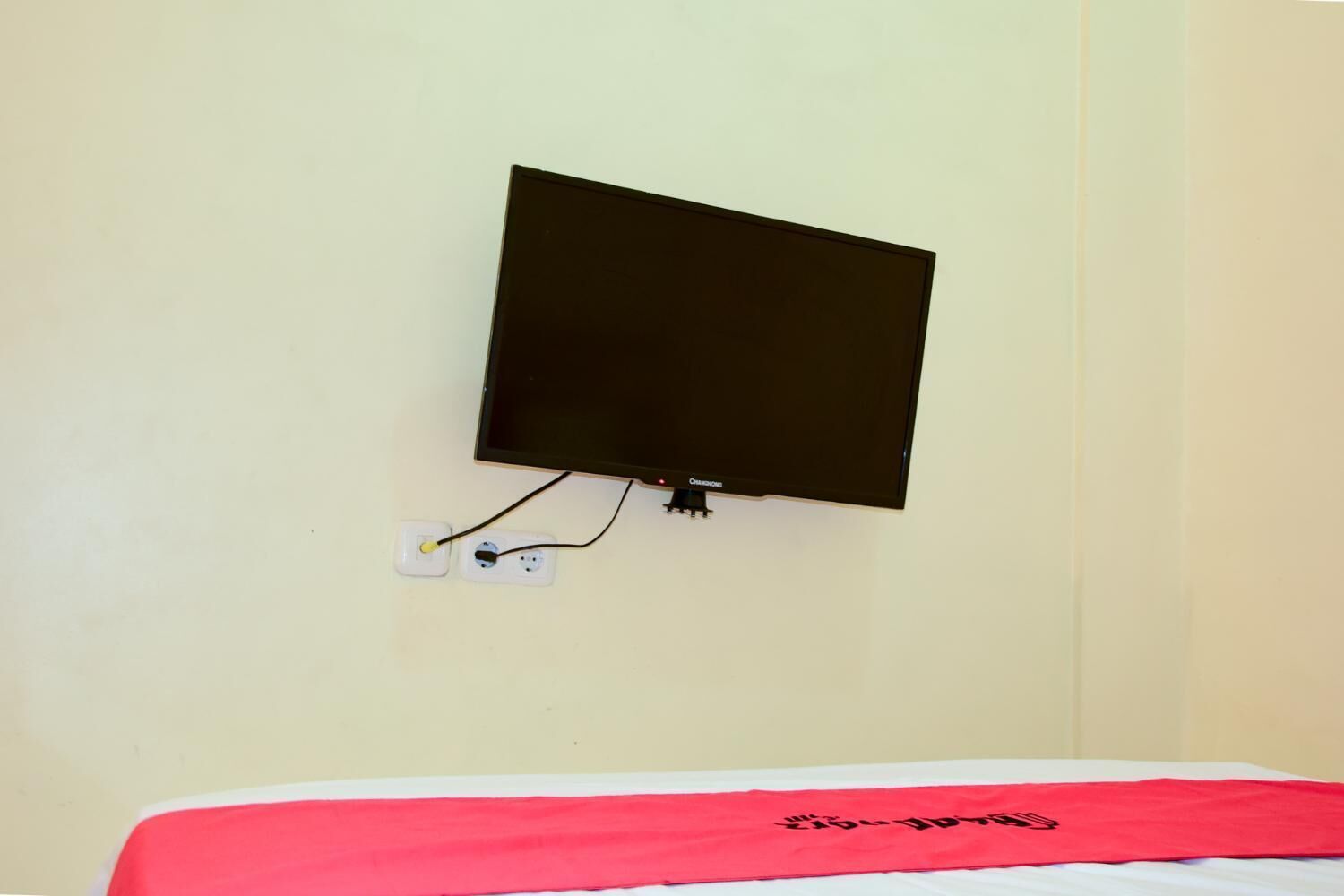 A bed with a red blanket and a television mounted on the wall.
