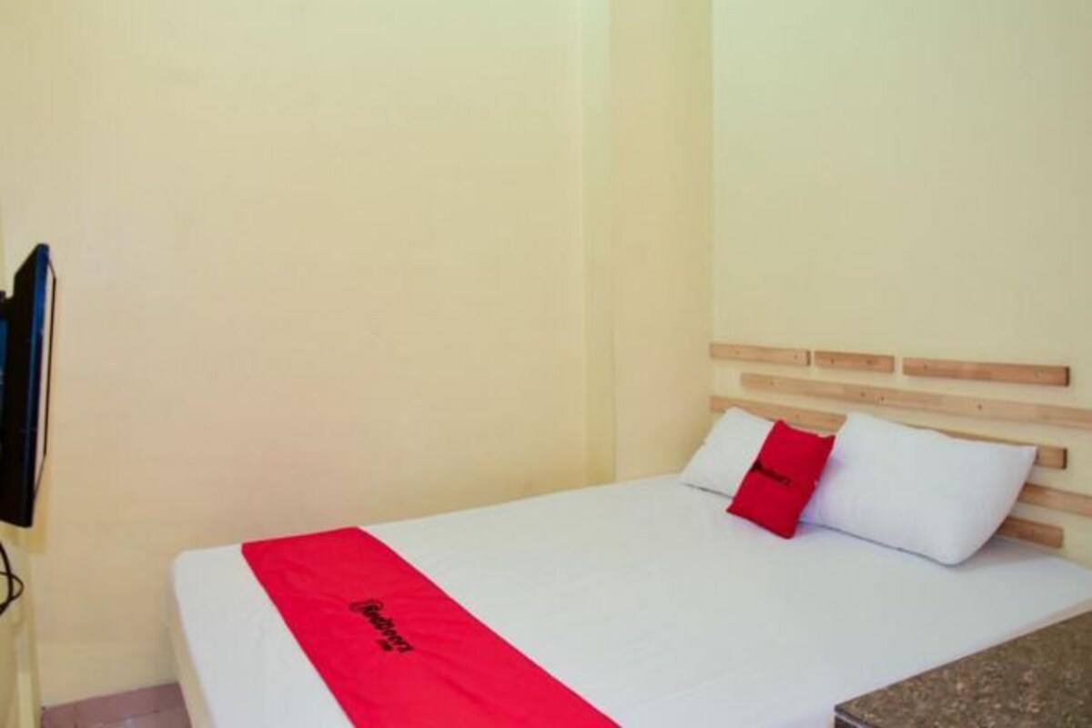 Un lit simple avec une tête de lit en bois, une couvre-lit blanche et rouge, et un oreiller rouge.