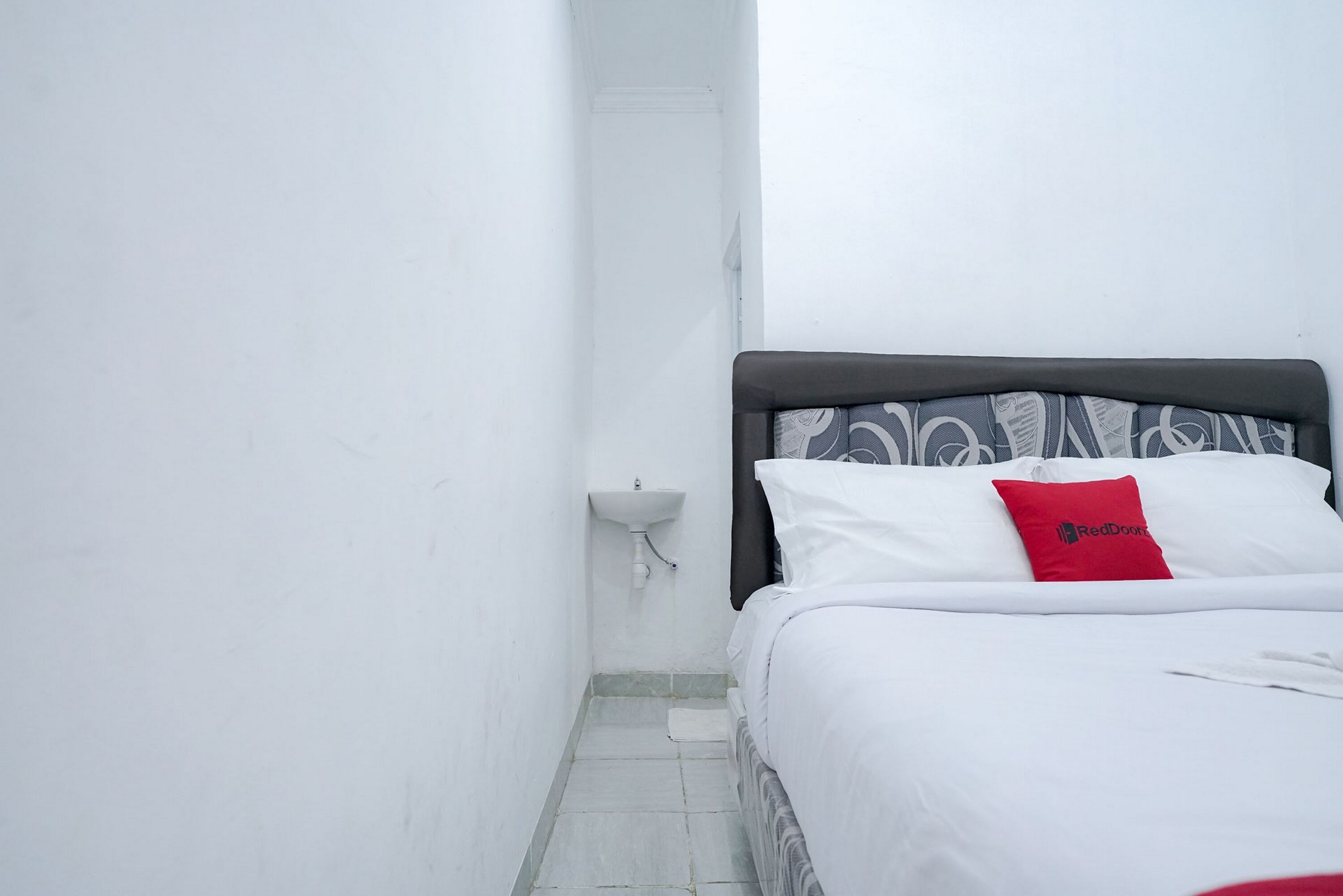A narrow hotel room with a bed, a bedside table, a small sink, and a red pillow.