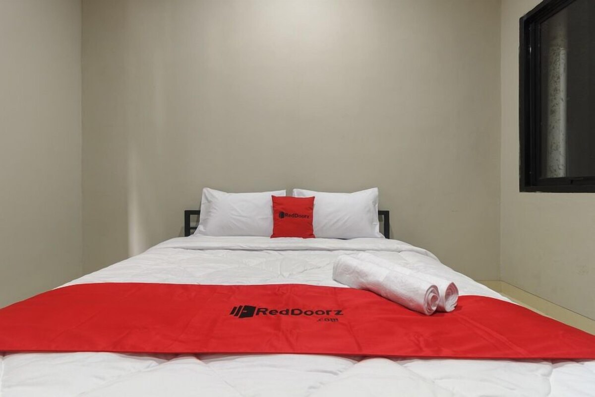 Un lit simple avec une literie blanche et une couverture rouge, un oreiller orné d’un logo rouge et une serviette enroulée posée sur le lit.
