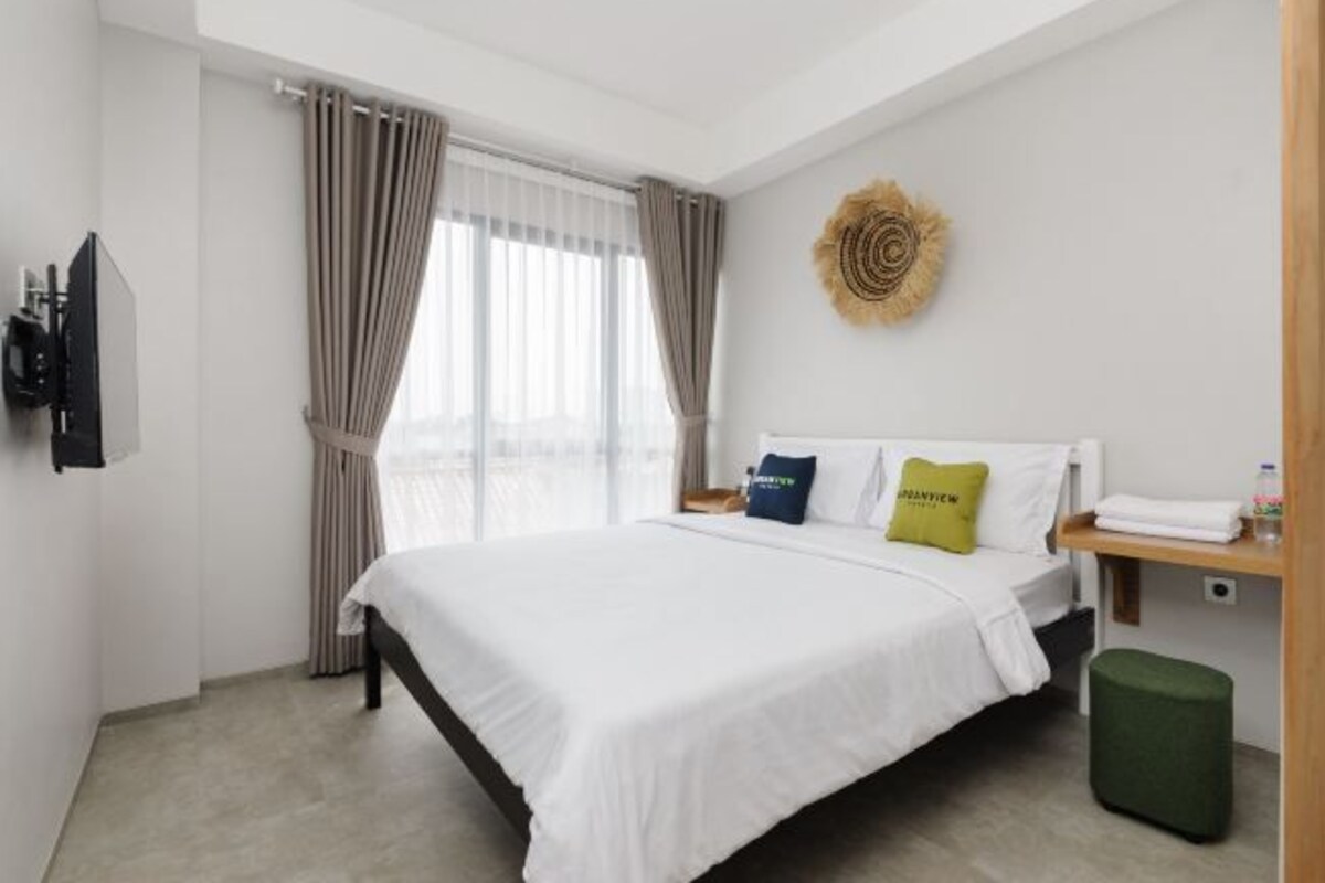 Un cuarto de hotel con una cama grande, un televisor de pantalla plana montado en la pared, una mesita de noche de madera, un reposapiés verde y un adorno de pared.