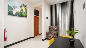 Un hall d’hôtel avec un tableau représentant des oiseaux, un extincteur, un mur à rayures, un fauteuil à motifs floraux et une table sur laquelle est posée une plante en pot.