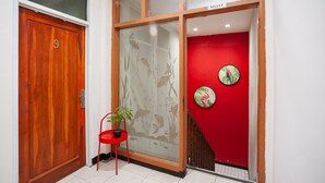 Une porte en bois avec une plaque portant un numéro, un mur rouge orné d’un motif circulaire et une chaise rouge sur laquelle est posée une plante en pot.