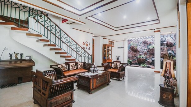 A spacious living area with a wooden staircase, leather sofas, and a large window overlooking a rocky garden. - RedDoorz Syariah near Gelora Delta Sidoarjo (Sidoarjo)