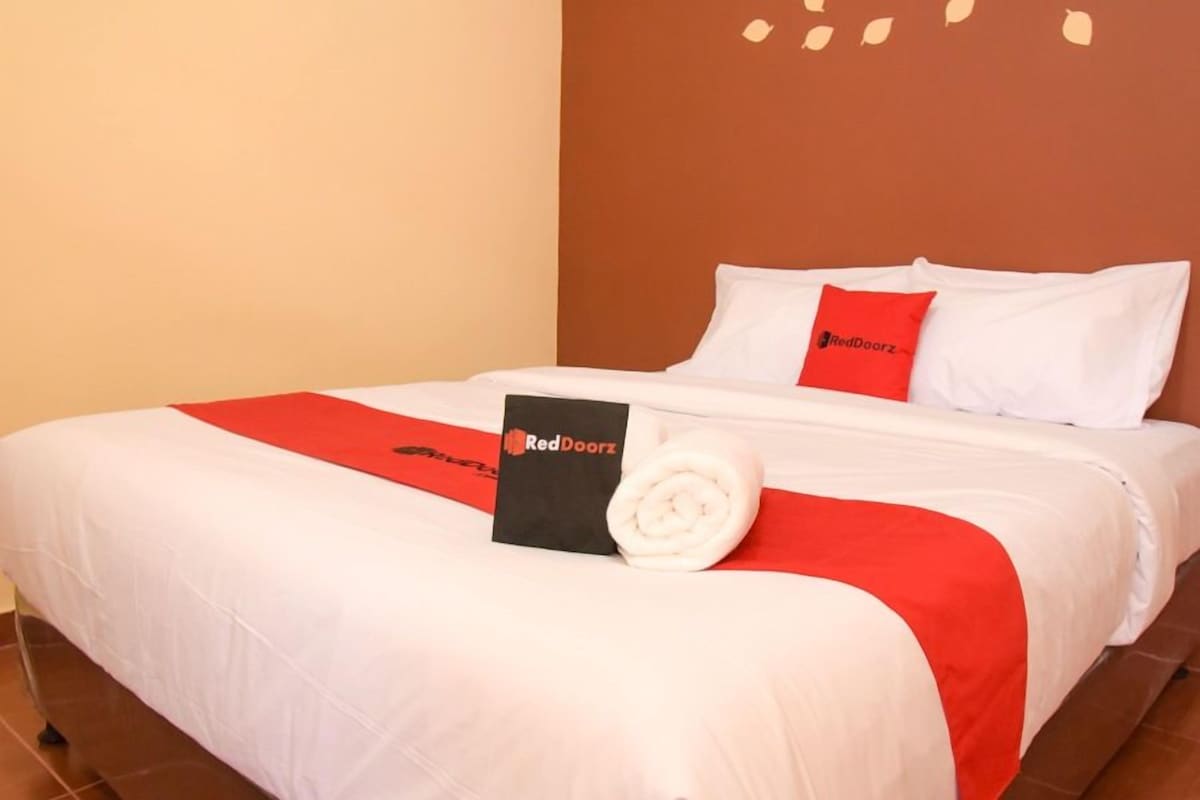 Una cama bien hecha con ropa de cama blanca y roja, una toalla enrollada y un letrero de RedDoorz sobre la cama.