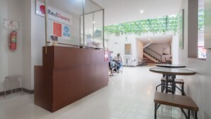 A reception area with a wooden desk, a fire extinguisher, and a sign for RedDoorz. There are tables and chairs in the background, and a staircase is visible. - RedDoorz Plus @ Dago Pojok 3 (Bandung)