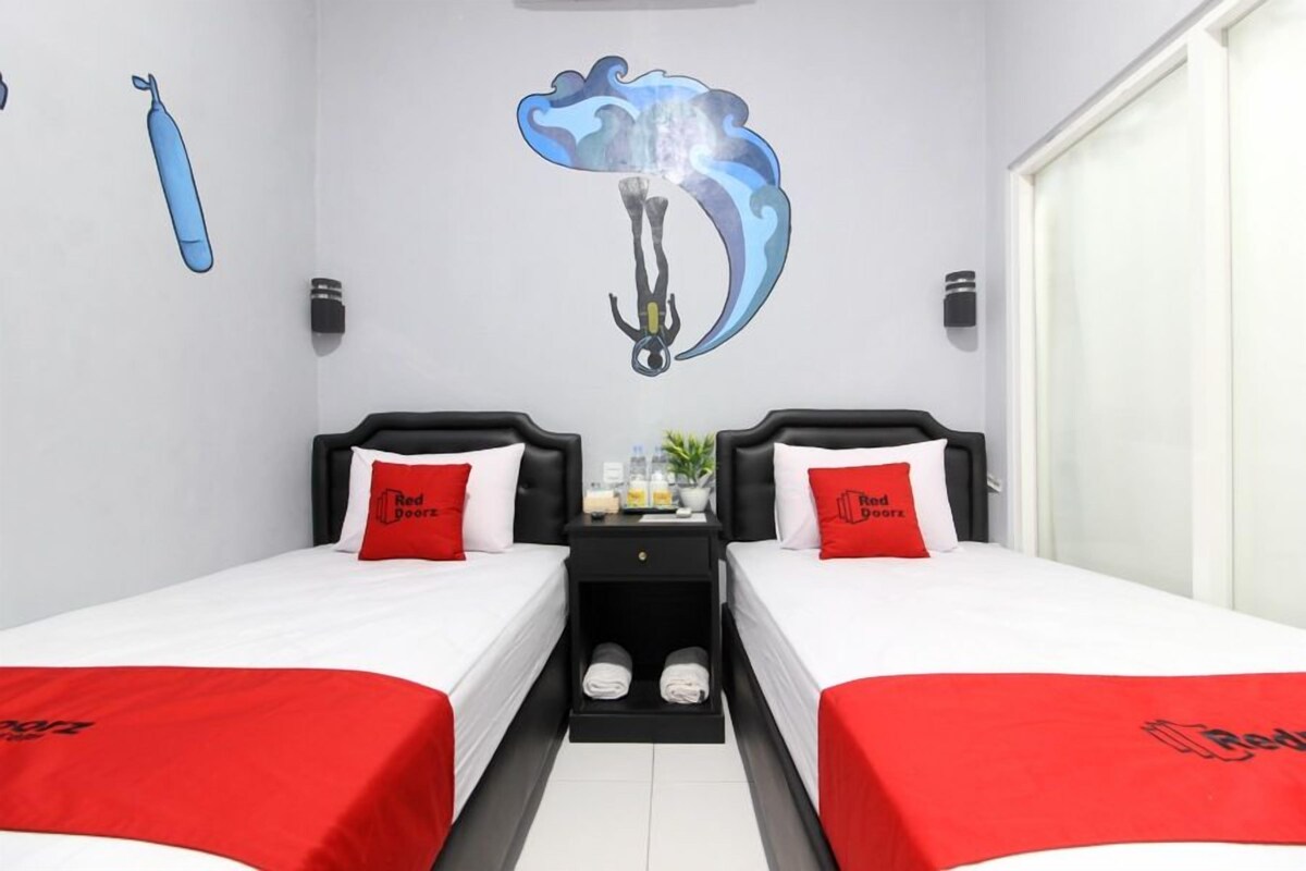 Quarto de hotel com duas camas, cada uma com um cobertor e travesseiro vermelhos, uma pequena mesa com uma planta, e um mural na parede.