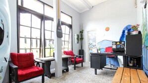 A room with large windows, a surfboard, red chairs, a wooden table, and a black speaker. - RedDoorz near Graha Saba UGM (Depok)