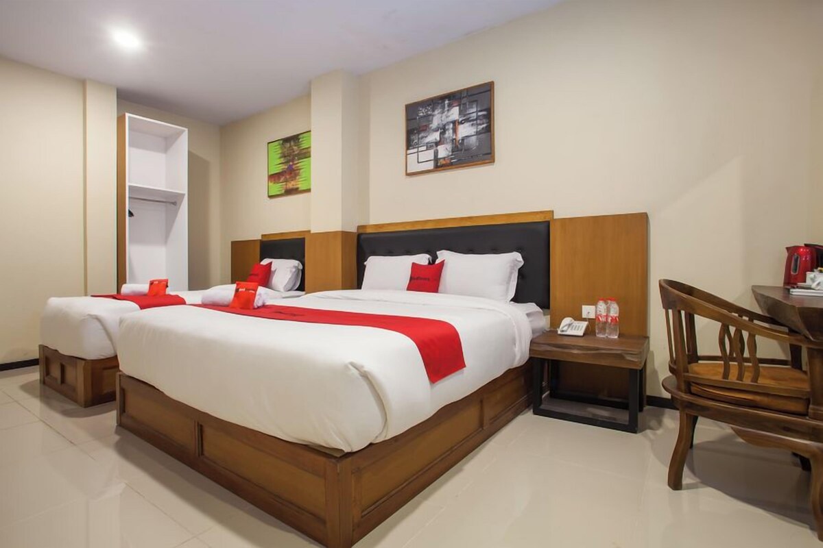 Habitación de hotel con una cama grande, dos mesitas de noche, una silla de madera y una pequeña mesa con una tetera roja.
