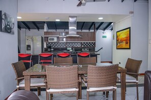 A modern kitchen with stainless steel appliances, a dining area with wicker chairs, and a view of the sea through a window.
