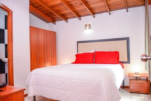 A bedroom with a wooden ceiling, a bed with white bedding and red pillows, a wooden wardrobe, and a bedside table with a lamp.
