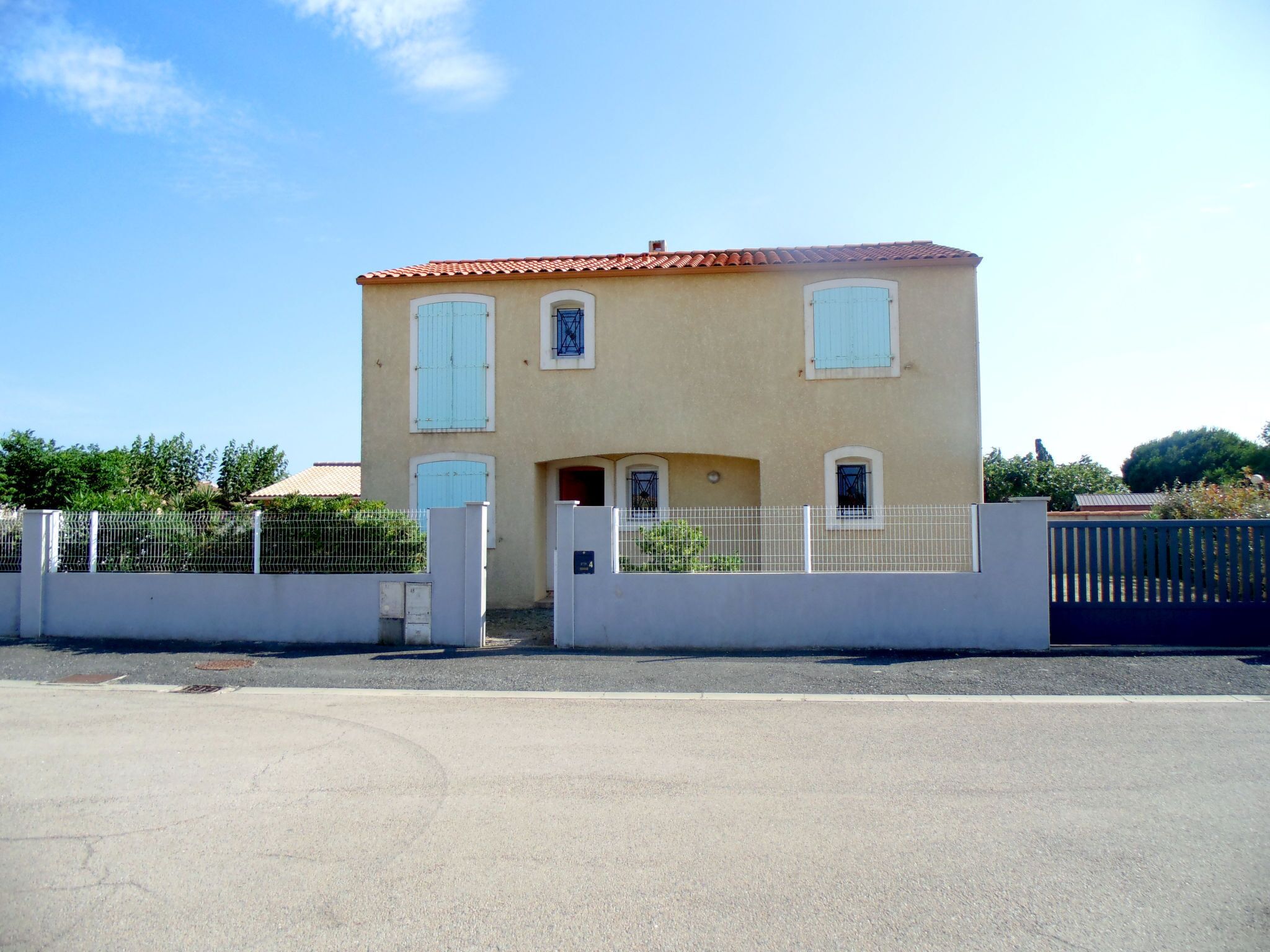 A two-story building with a beige facade and red-tiled roof. It features a gated entrance with a metal fence and a small garden area in front.