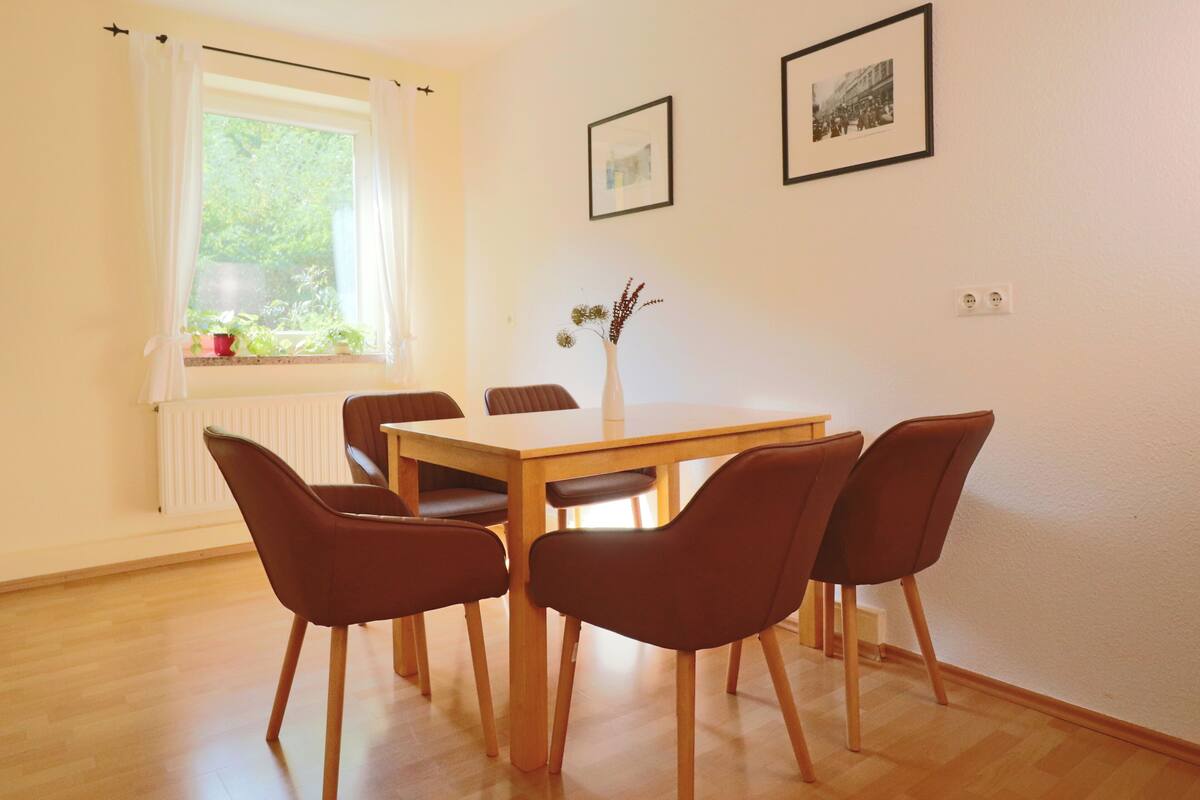 Un comedor con una mesa de madera y cuatro sillas marrones, un florero con flores y dos cuadros en la pared.
