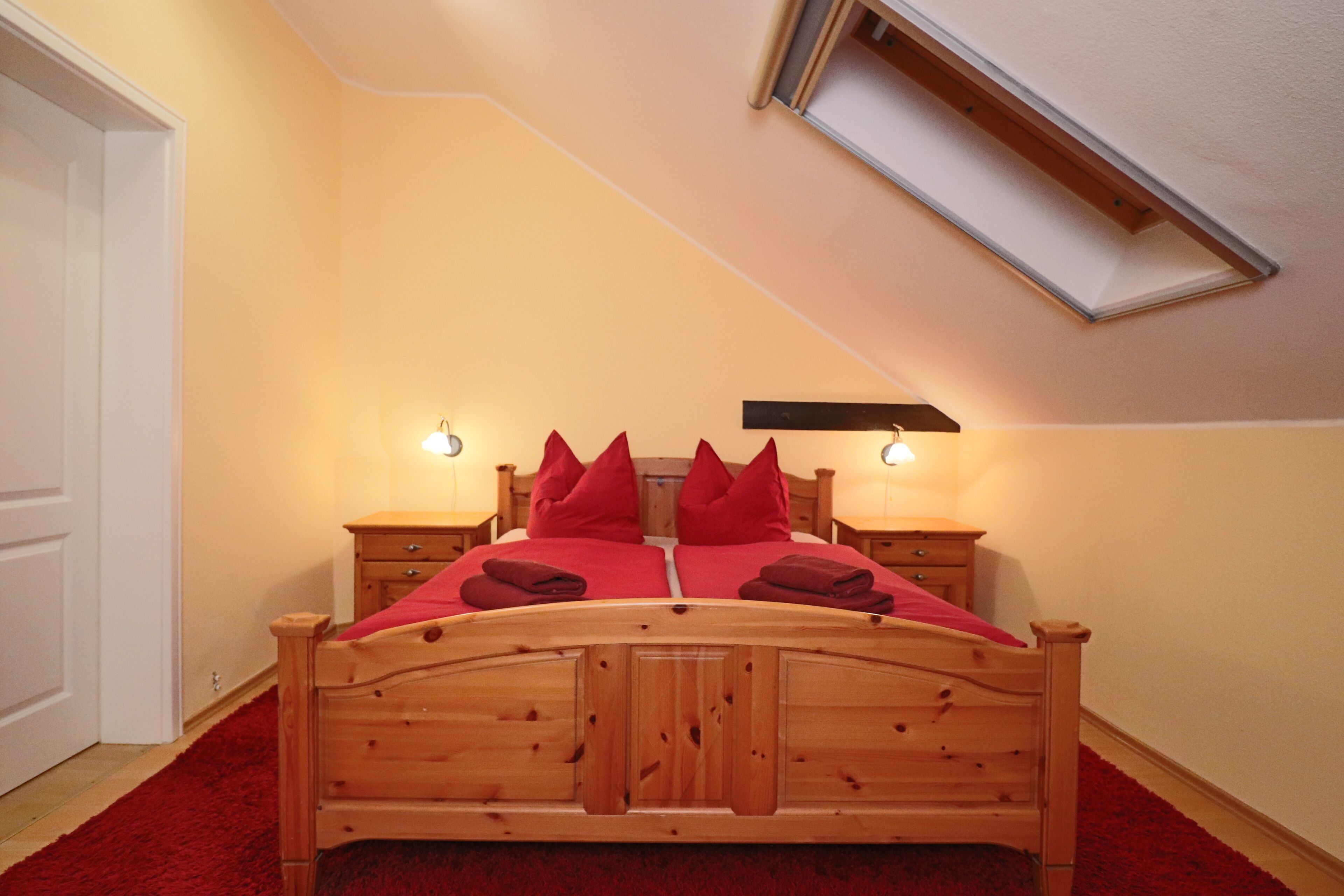 A bedroom with a wooden bed, red bedding, two bedside tables, a skylight, and a red carpet.