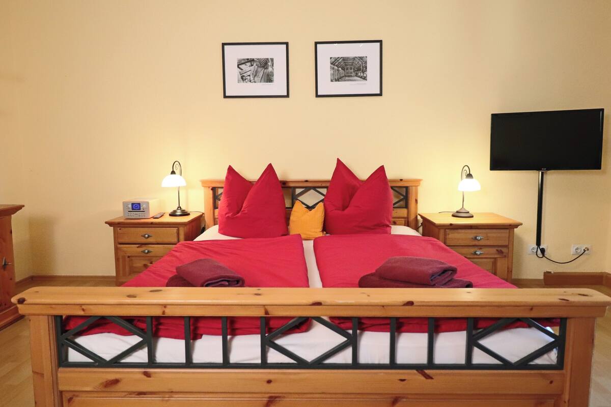 Una cama bien hecha con almohadas rojas y amarillas, dos mesitas de noche con lámparas y un televisor de pantalla plana montado en la pared.
