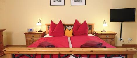 Una cama bien hecha con almohadas rojas y amarillas, dos mesitas de noche con lámparas y un televisor de pantalla plana montado en la pared.